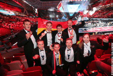 acht Menschen im Anzug mit Medaille, die eine Deutschlandfahne heben, in einer rot beleuchteten Arena