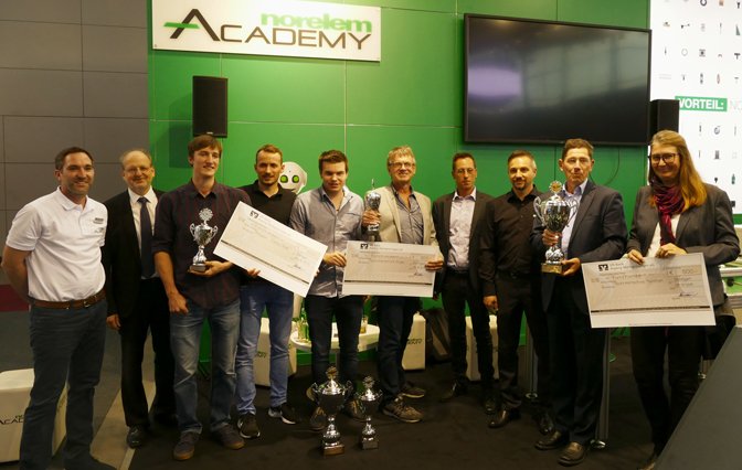 Engineering Newcomer Gewinner mit Pokal und Preisgeld Scheck in der Hand vor einer grau grünen Werbetafel mit Fernsehgerät